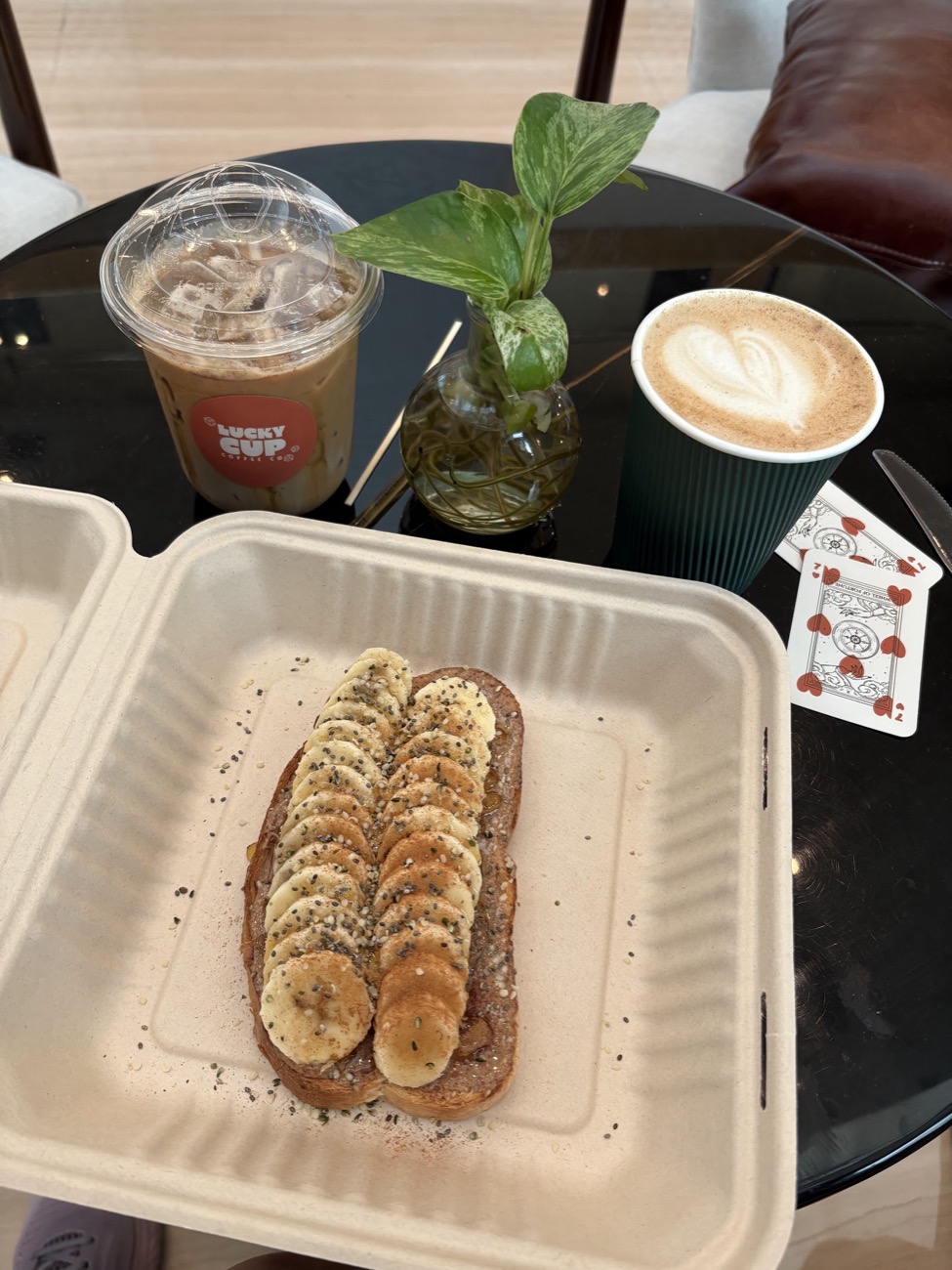 Banana toast topped with sliced banana and seeds in a takeout box, beside an iced coffee, a hot latte and a sprig of greenery