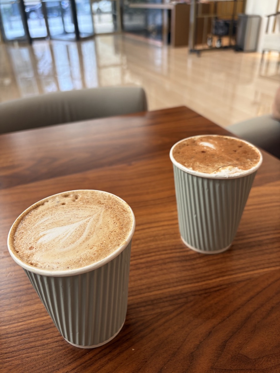 Two hot lattes in fluted cups on a wood table, one with a leaf poured into the foam
