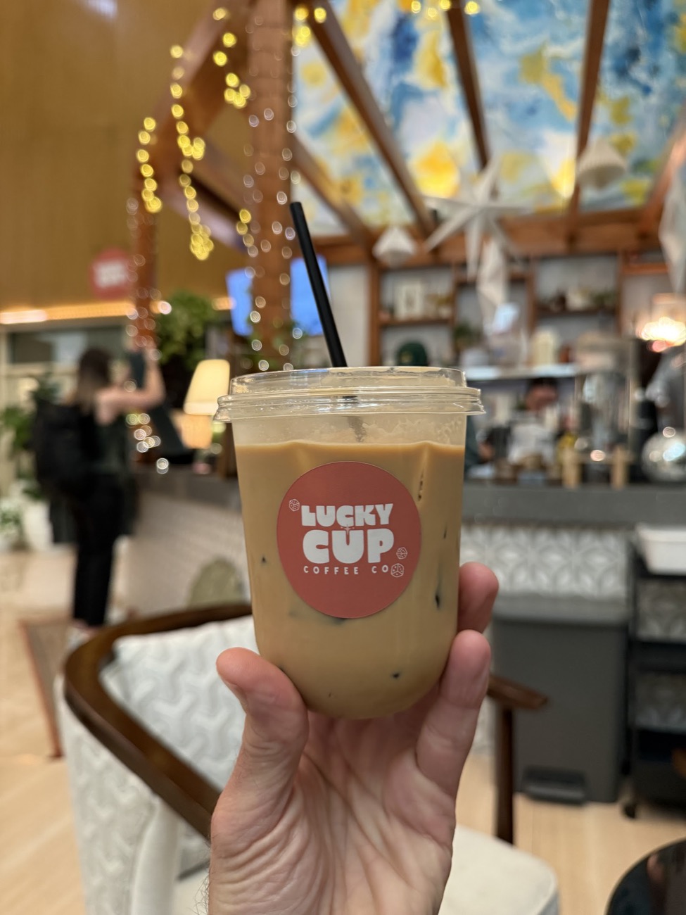 A hand holding an iced latte in a clear cup with the coral Lucky Cup sticker, in the bright hotel lobby