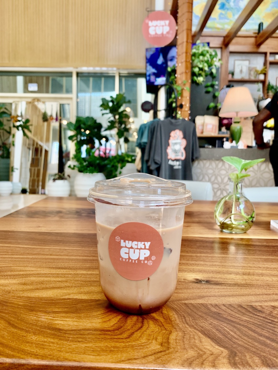 An iced latte with the Lucky Cup sticker on a wood table, with plants and a shelf of merchandise behind