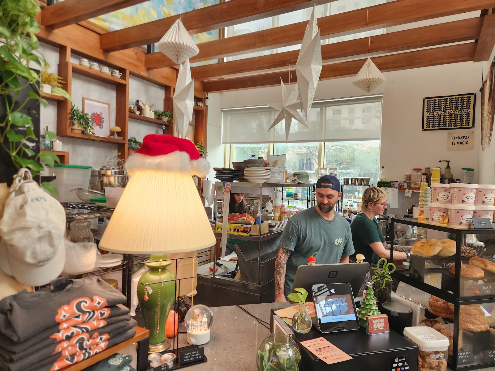 Inside Lucky Cup: a barista smiling behind the counter under hanging paper stars, with shelves of mugs, plants and a pastry case in the window light