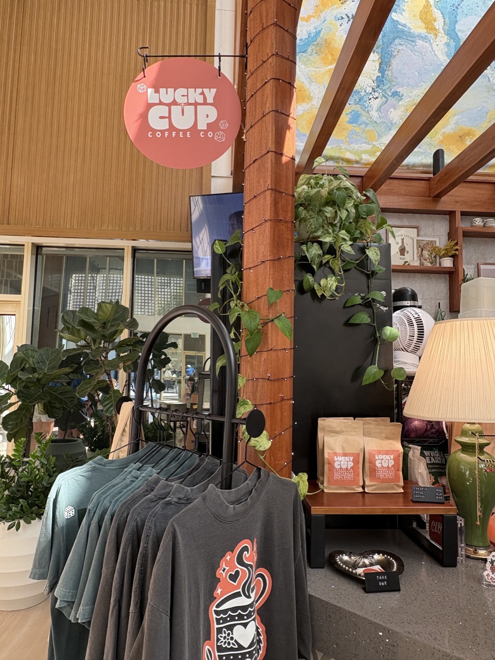 The round coral Lucky Cup Coffee Co. sign hanging by a wood post, above a rack of t-shirts and bags of coffee beans